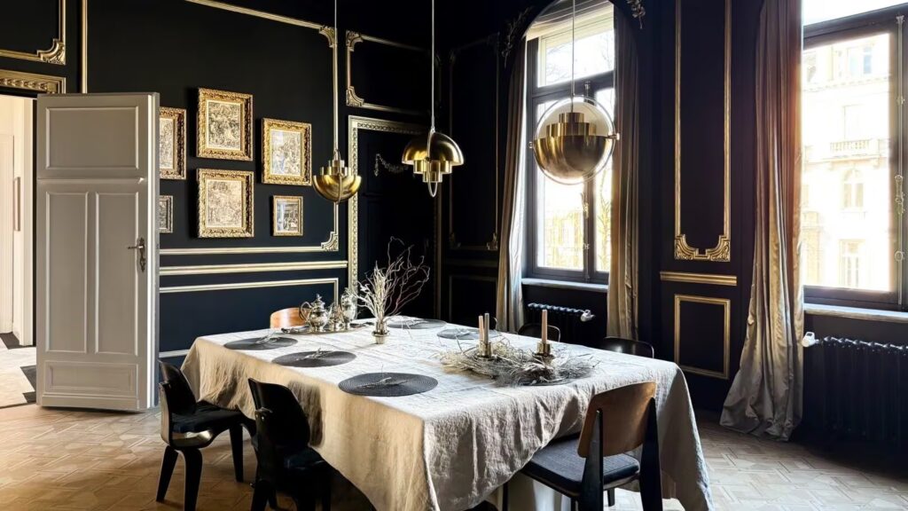 An opulent dining room in a historic mansion, featuring deep black walls with ornate gold leaf boiserie, a gallery wall of framed botanical and landscape art in gold frames, and multi-layered gold spherical pendant lights. A dining table with a linen cloth and centerpiece is set with dark chairs on a parquet floor, looking out over a large window to a city view.