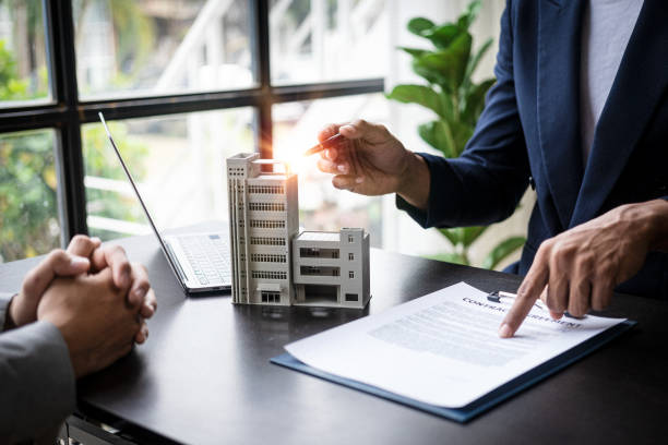 A professional Estate Manager in a blue blazer pointing to a contract agreement on a clipboard while discussing a multi-story architectural building model with a client. A laptop and greenery are visible in the bright, modern office background.