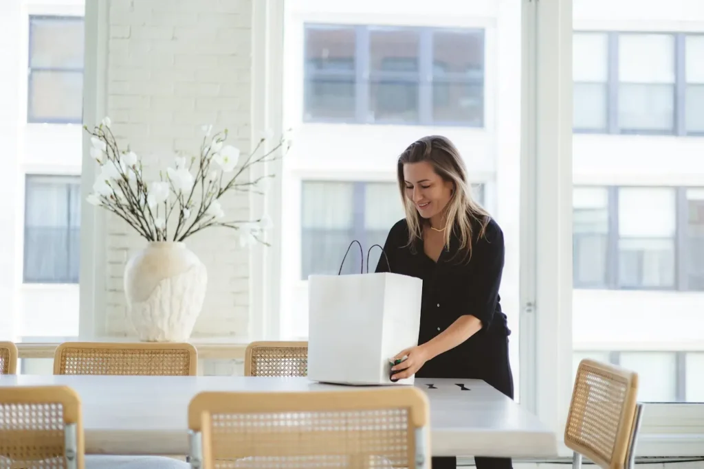 A professional woman in a black shirt smiling while organizing a white shopping bag on a light-colored dining table. The setting is a bright, modern penthouse or office with large windows, cane-back chairs, and a large vase of white flowers in the background.